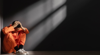 A distressed man dressed in an orange jumpsuit sitting on a jail floor with his hands on his head